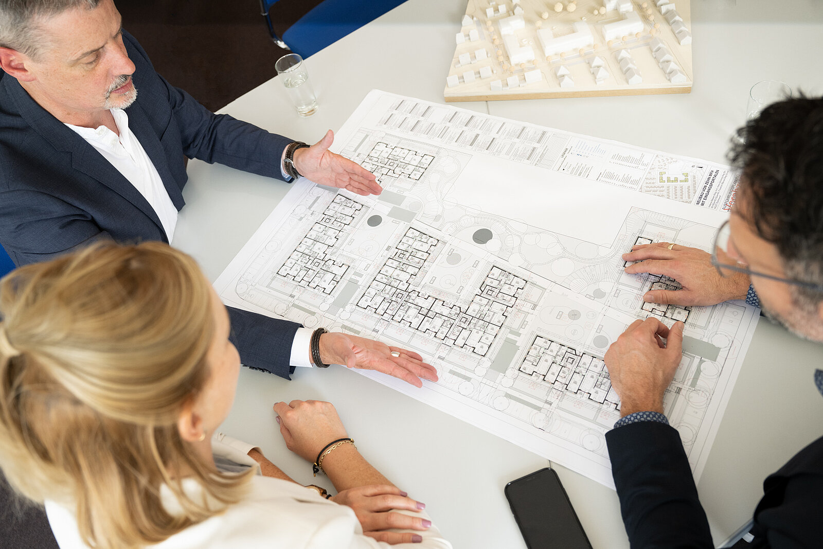 Ein Bauplan liegt auf einem Tisch. Ein Mann zeigt seiner Kollegin und seinem Kollegen etwas auf dem Plan. 